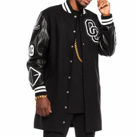 Model wearing a long black varsity coat with sleeve patches, snap buttons and striped cuffs.