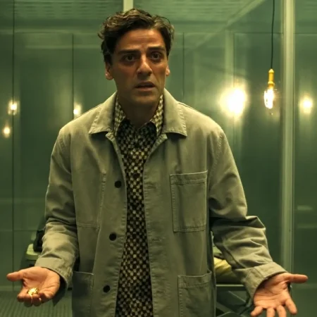 Man wearing a grey chore jacket over a patterned shirt in a green-lit mirrored room with hanging light bulbs.