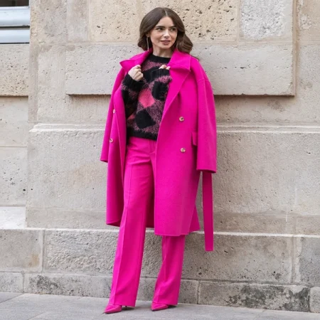 Woman wearing a hot pink coat over a black and pink sweater with matching pink trousers beside a stone wall.